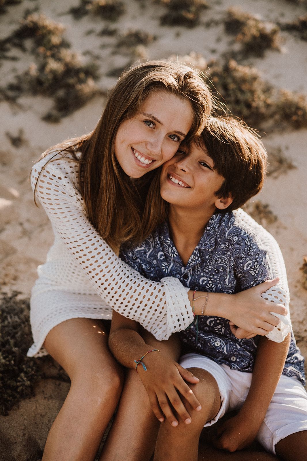 A smiling mother in a white sweater tenderly embraces her young son on a sandy beach.