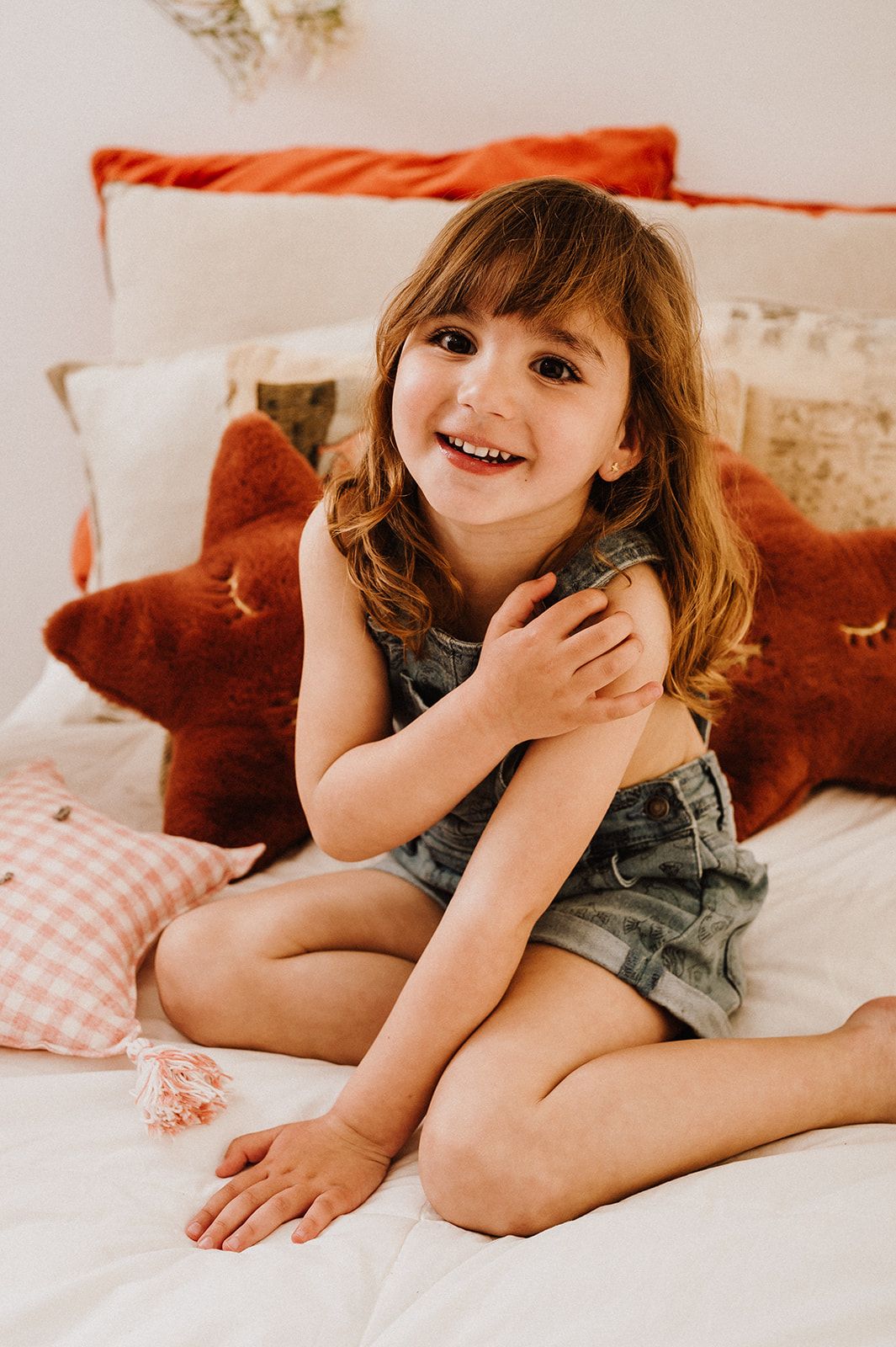 Smiling young girl in denim overalls sitting cross-legged on a bed surrounded by colorful pillows.
