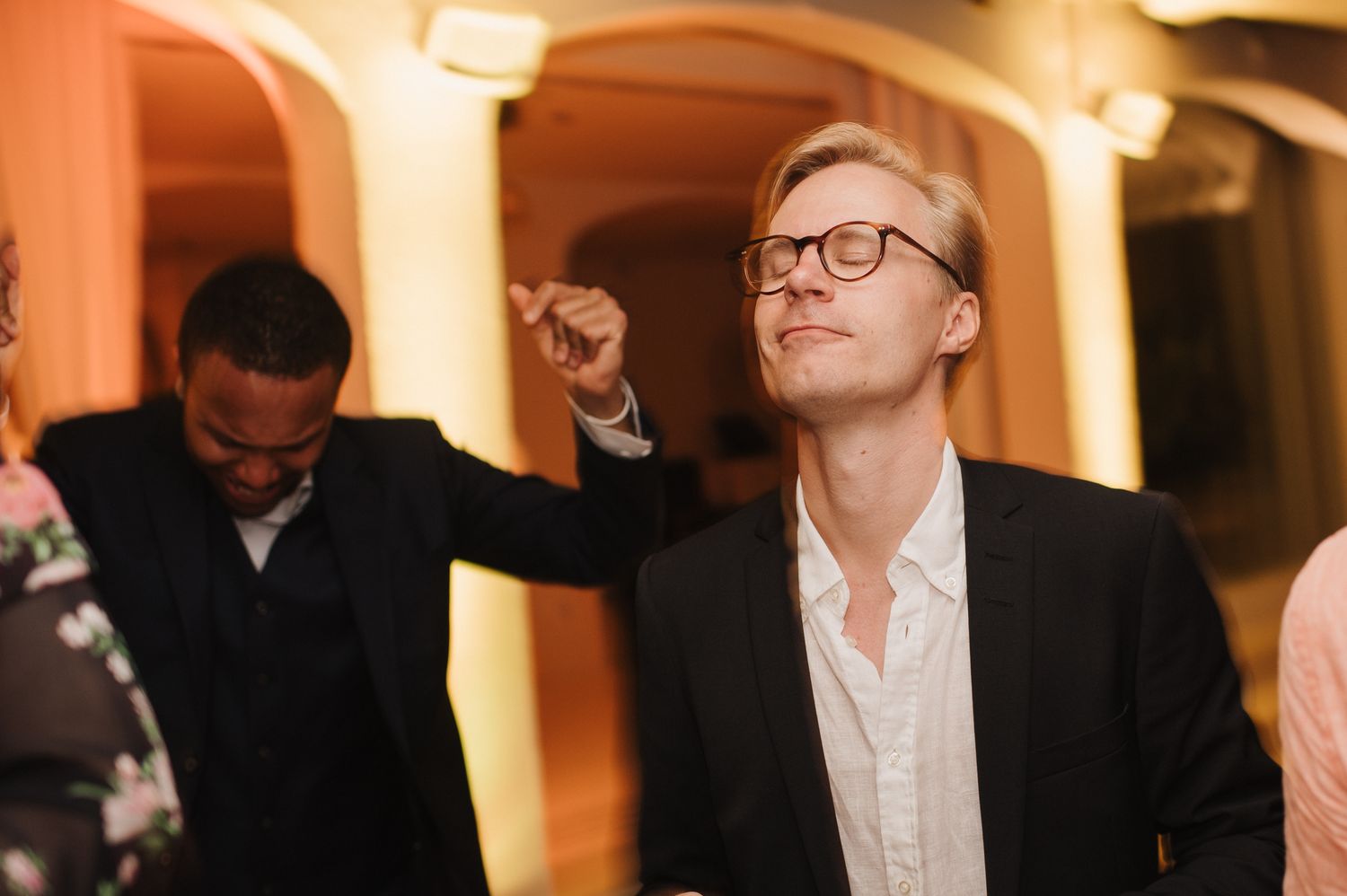 Smiling man in glasses points upward while dancing at an indoor wedding reception party.