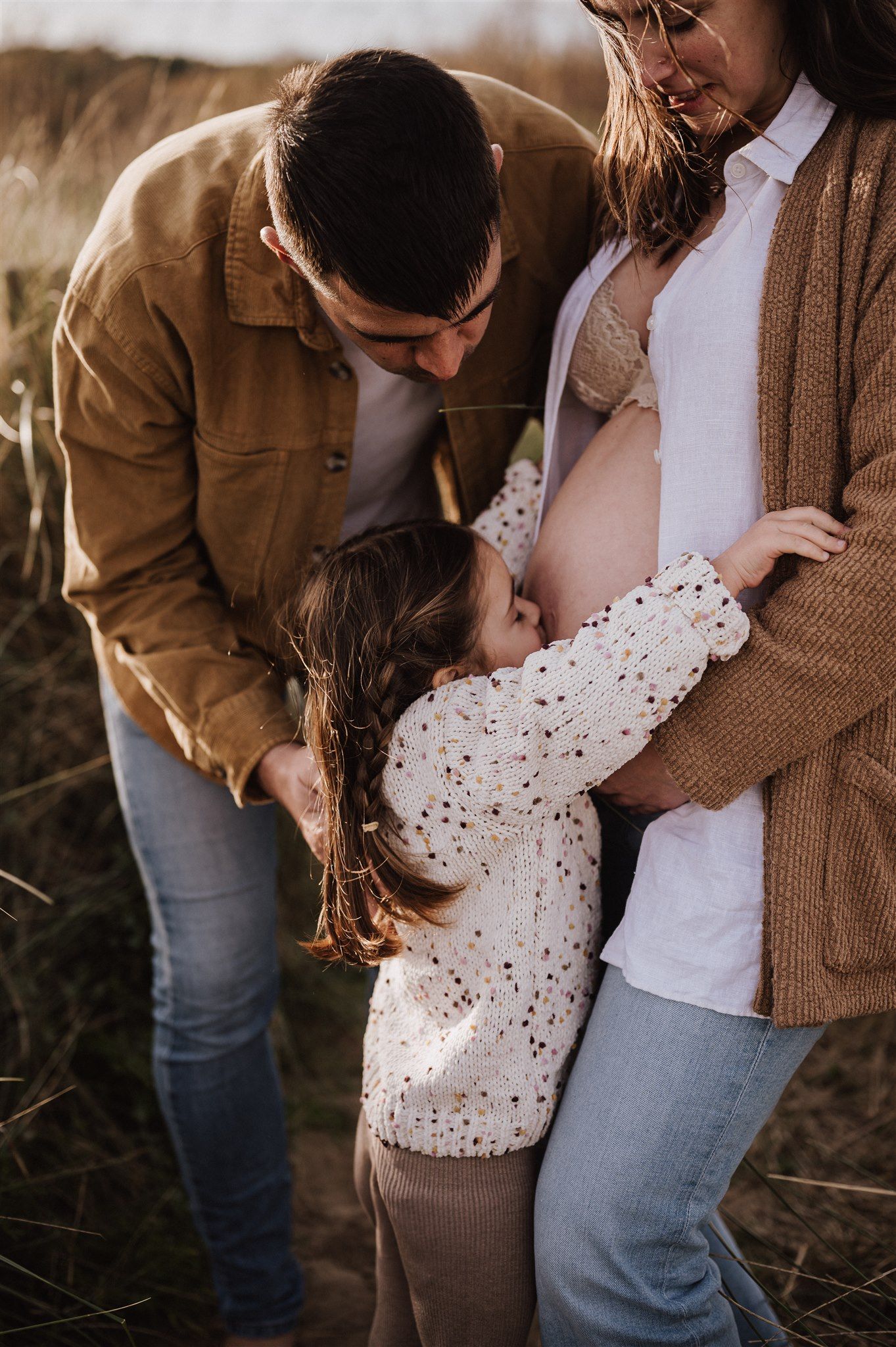 A family tenderly embraces a pregnant mother outdoors, with a young girl kissing her baby bump in a warm autumn setting.