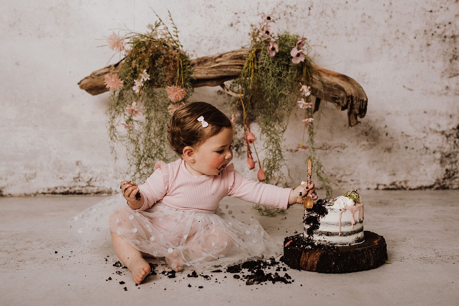 Toddler girl in pink tutu smashing first birthday cake on wooden stump with floral swing backdrop.