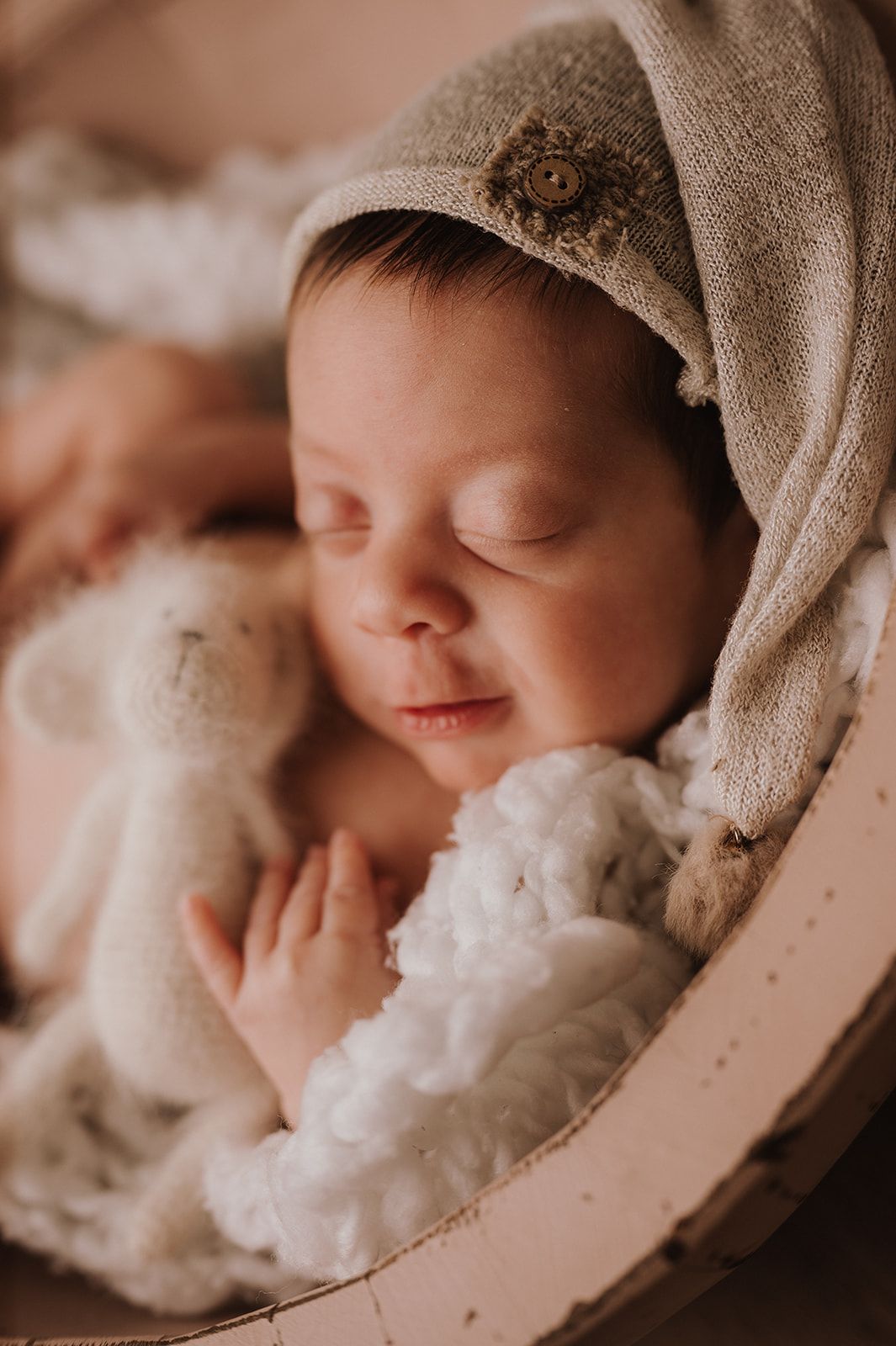 Peaceful newborn baby sleeping in a round basket with a soft teddy bear and draped linen bonnet.