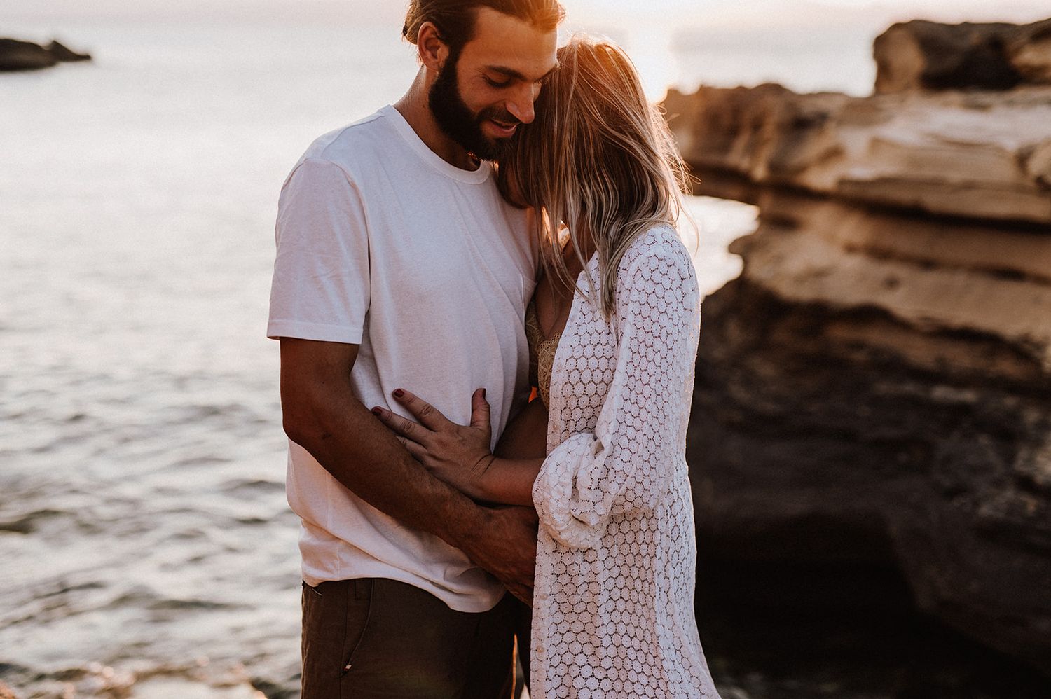 Couple embracing tenderly on rocky beach at golden hour with ocean backdrop.