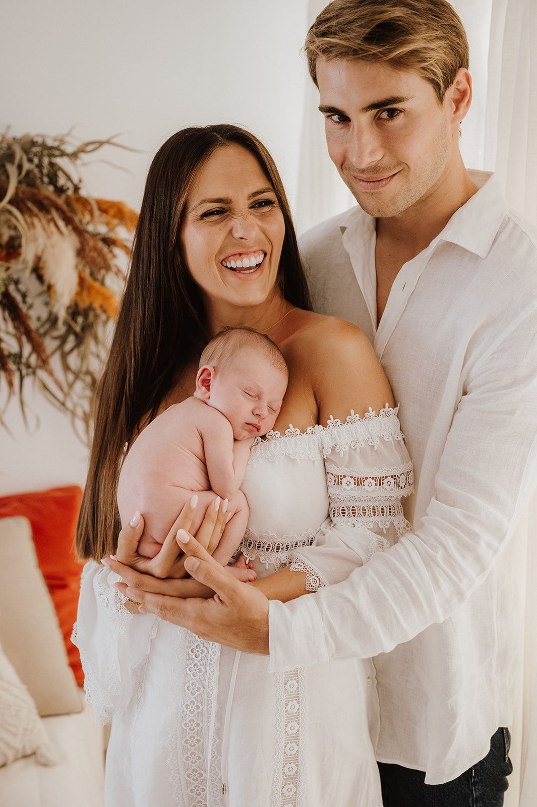 Happy new parents in white outfits smiling and holding their sleeping newborn baby in a bright room.