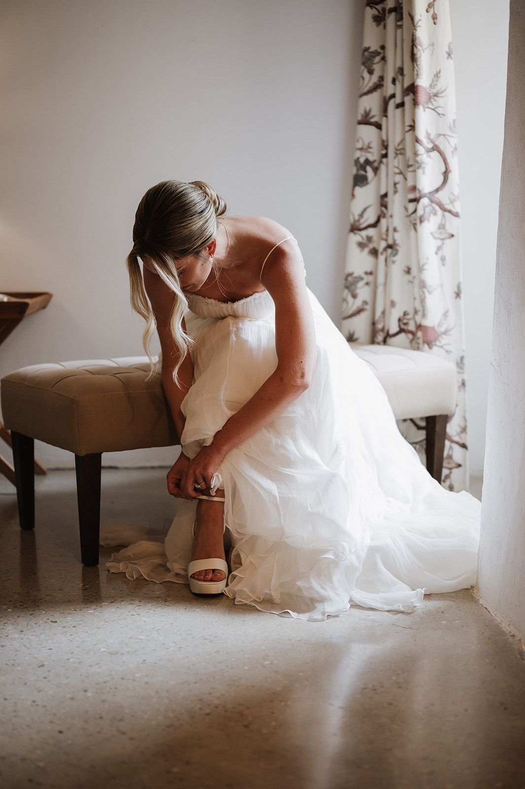 Bride in white gown fastening her wedding shoes while seated on a bench near a floral curtained window.