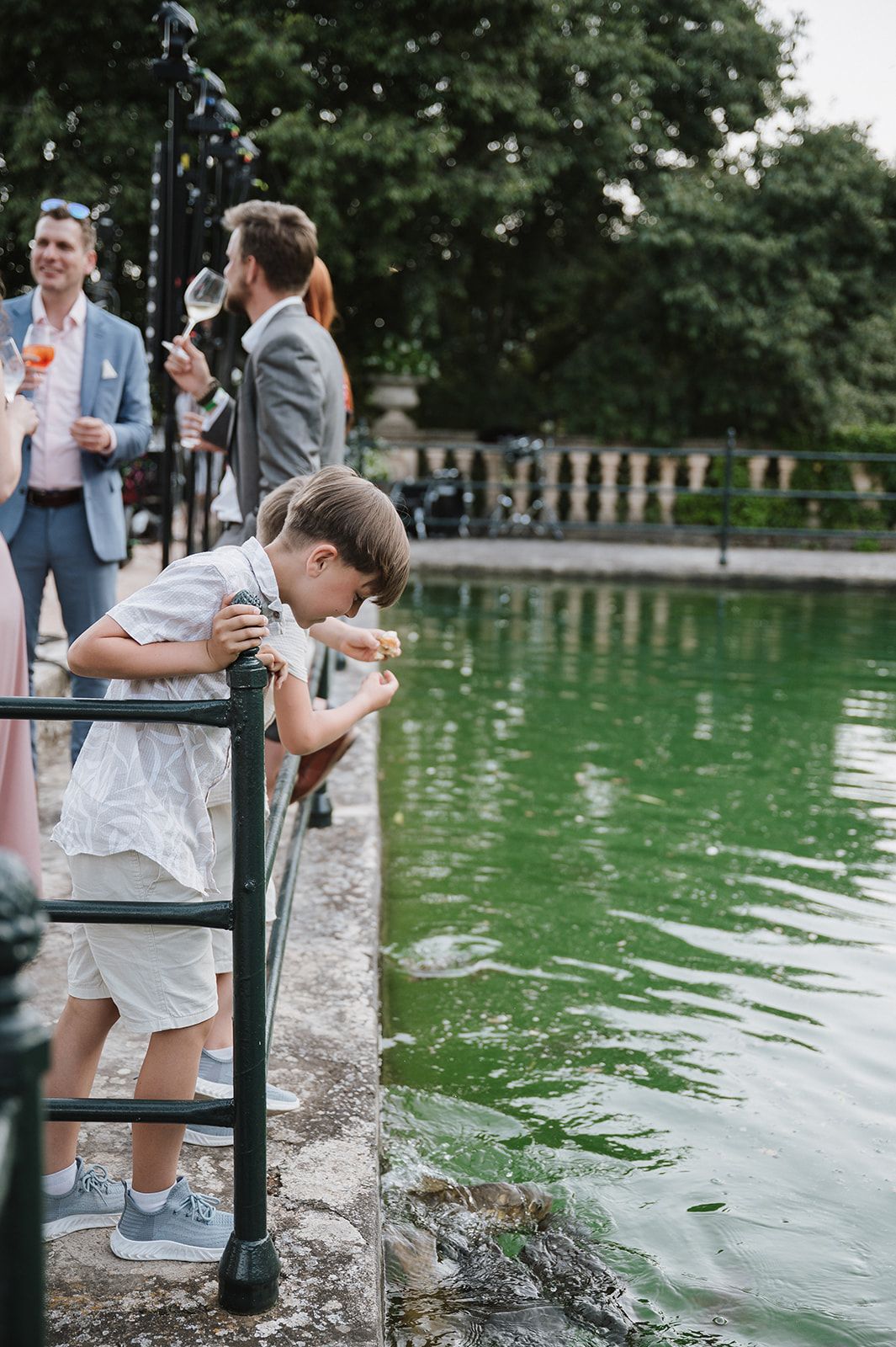 Young boy in white outfit leans over railing feeding ducks in green pond at outdoor wedding.