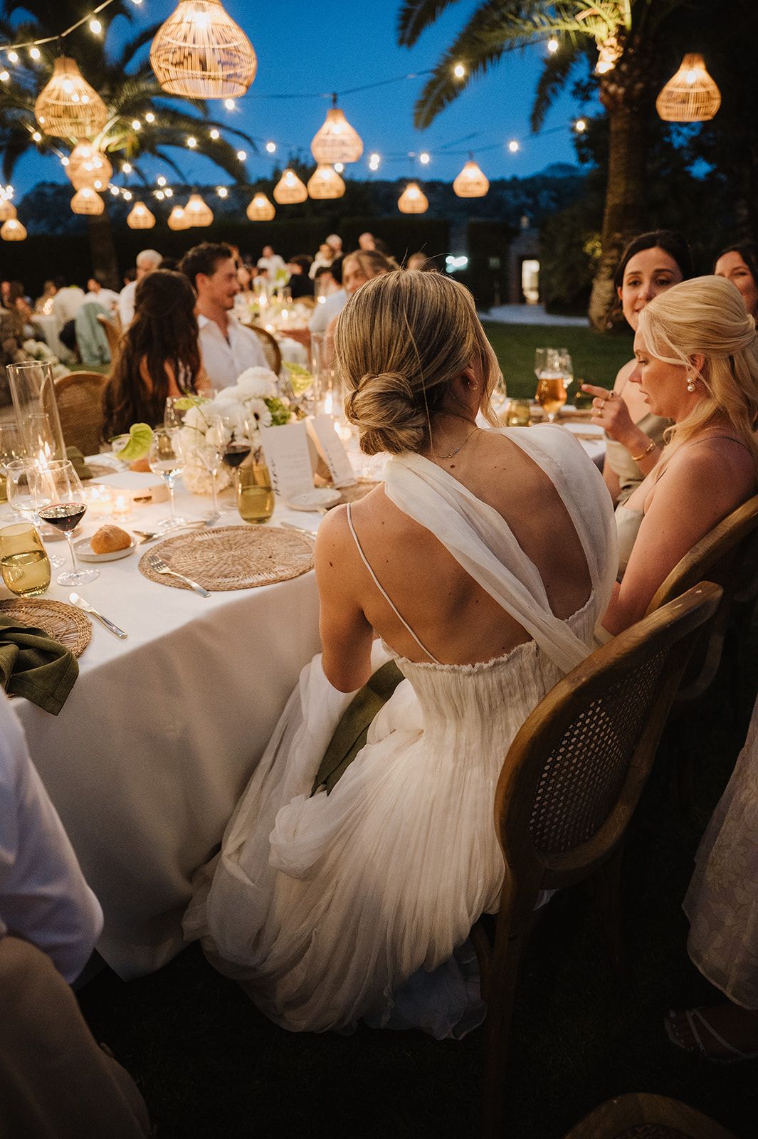 Bride in backless white gown seated at candlelit outdoor wedding reception under glowing pendant lights.