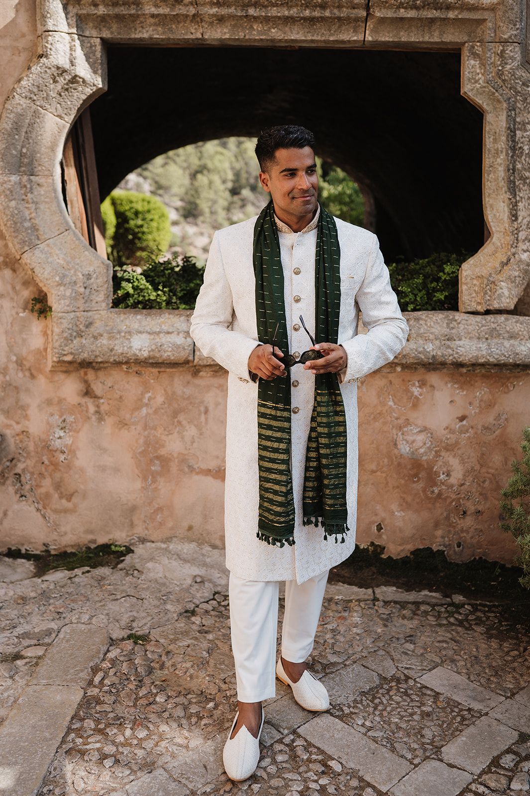 Groom in white sherwani with green dupatta scarf at outdoor Indian wedding ceremony.
