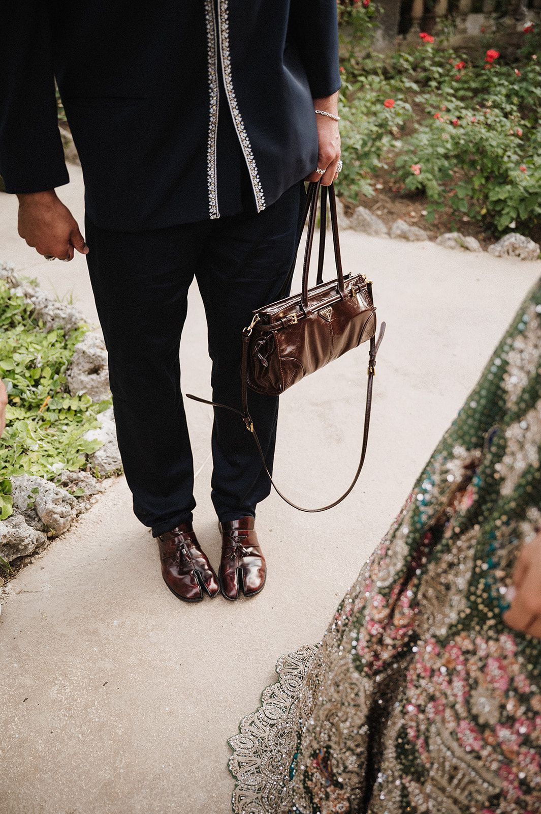 Groom in black kurta holding bride's embellished bag and wearing brown leather shoes outdoors.
