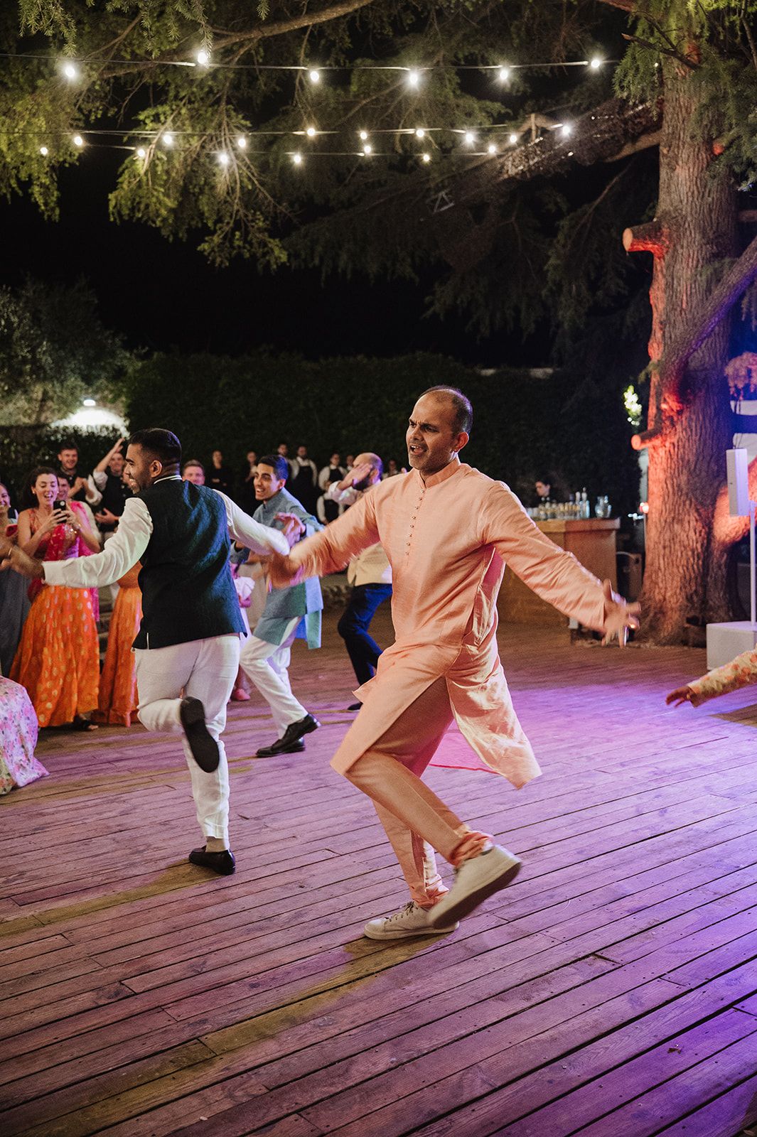 Guests dance joyfully on outdoor wooden dance floor at night during a vibrant Indian wedding celebration.