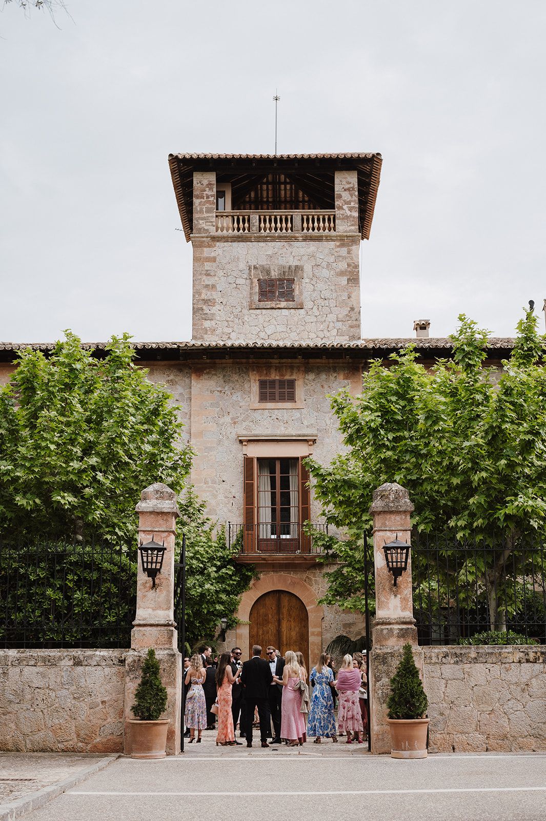 Wedding guests gather outside rustic stone villa with tower entrance framed by manicured trees and iron gates.
