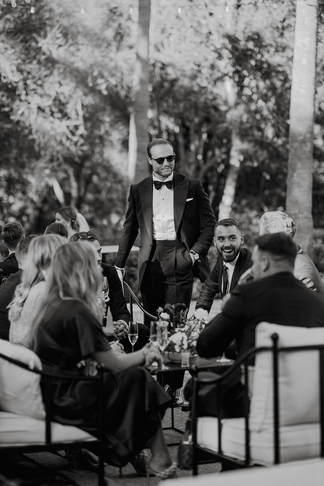 Groom in tuxedo and sunglasses smiles while greeting seated wedding guests at outdoor reception.