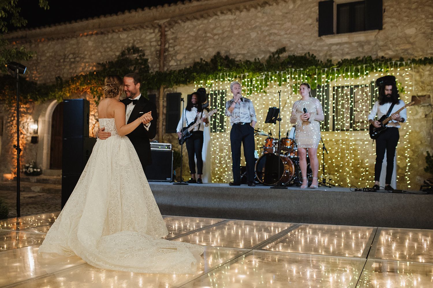 Bride and groom share first dance on illuminated floor with live band performing behind them.