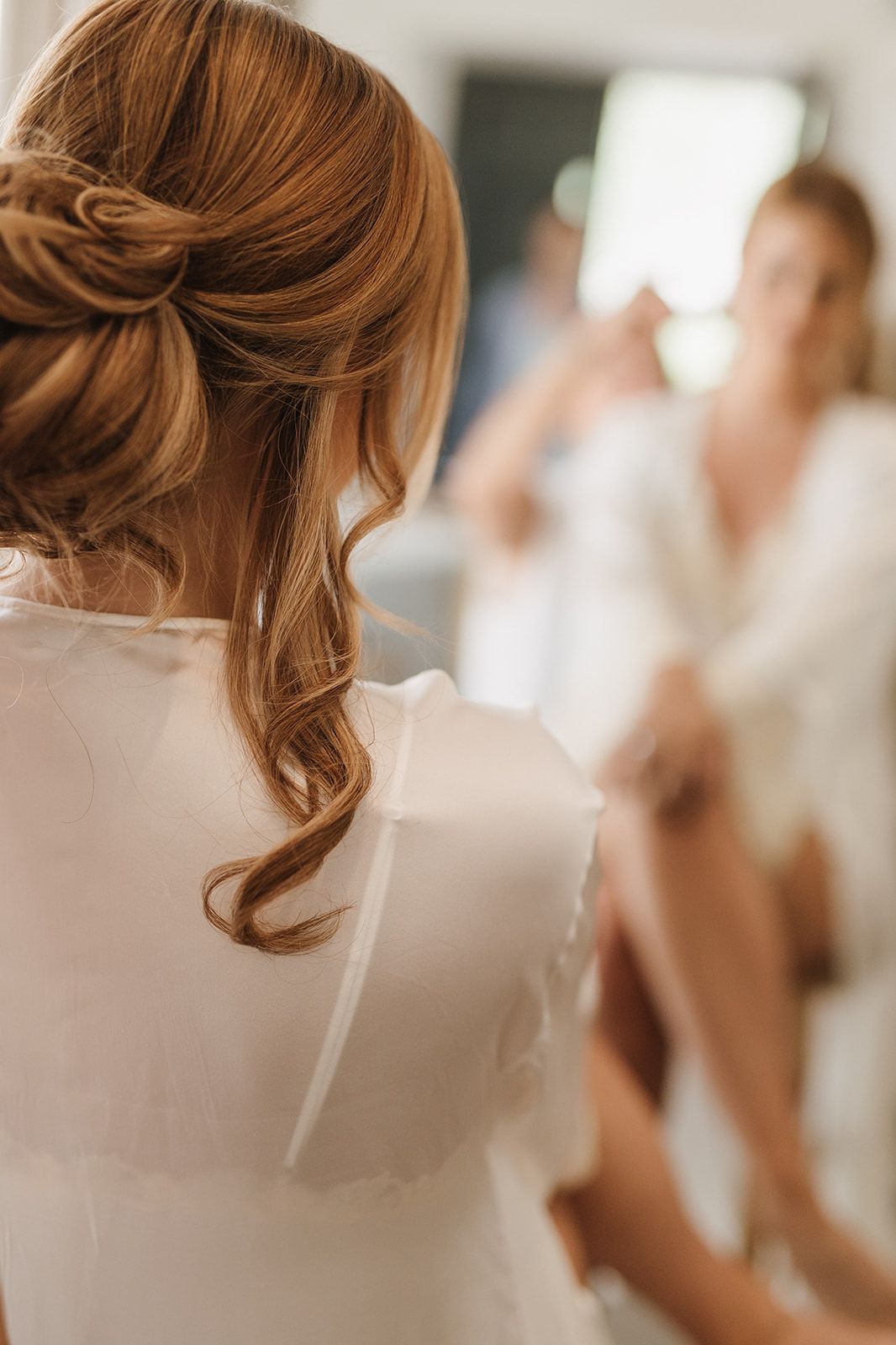 Bride with elegant half-up auburn hairstyle in white robe preparing for wedding day with bridesmaid in background.