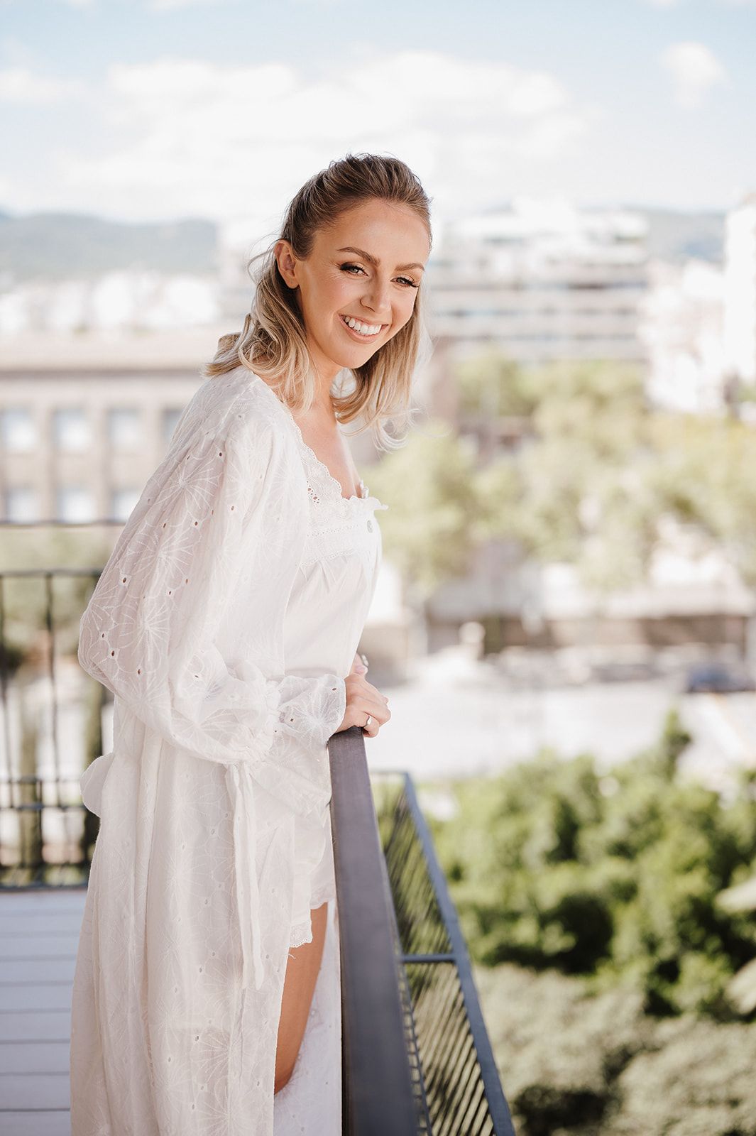 Smiling blonde woman in white lace robe on a sunny balcony with city views.