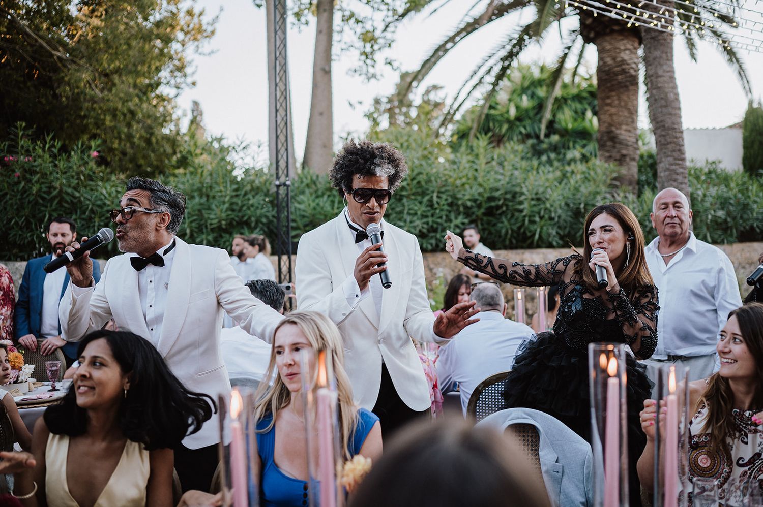 Man in white tuxedo giving speech at elegant outdoor wedding reception with guests seated at decorated tables.