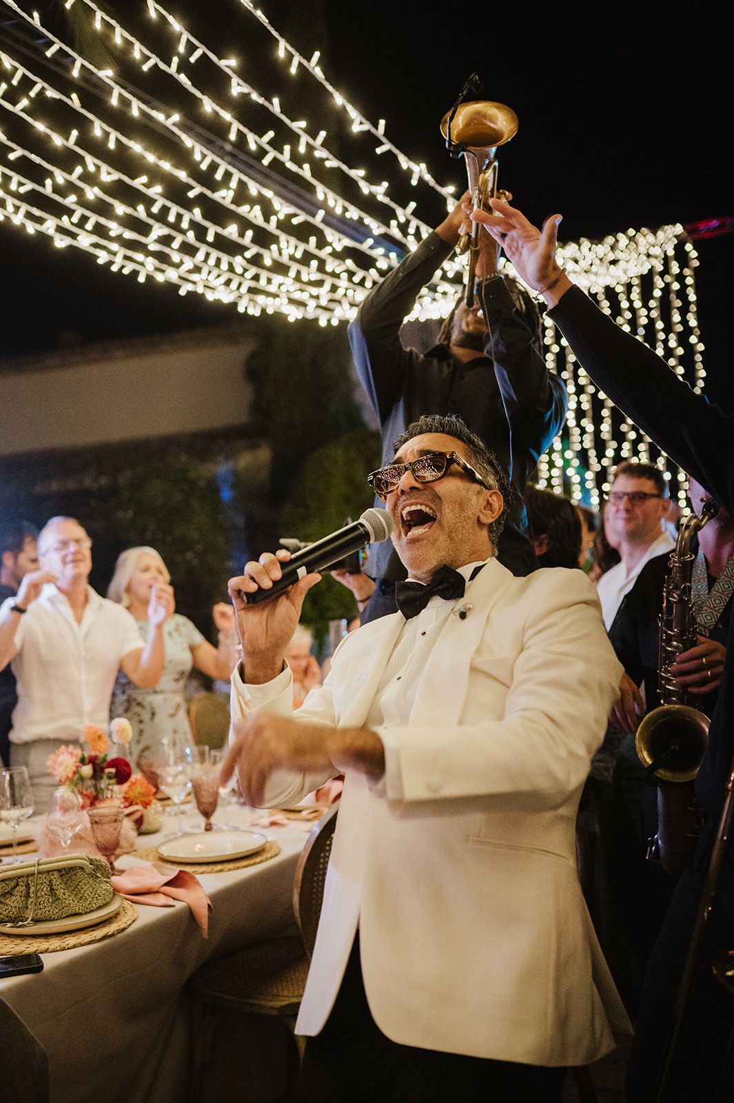 Energetic wedding singer in white tuxedo performs with microphone while band plays trumpet and saxophone at reception.
