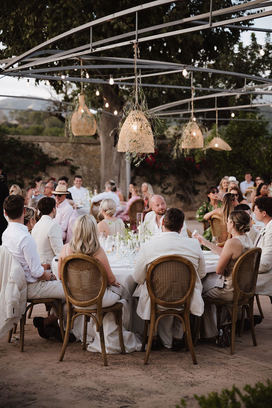 Guests seated at elegant round tables under string lights and woven pendant lamps at an outdoor wedding reception.
