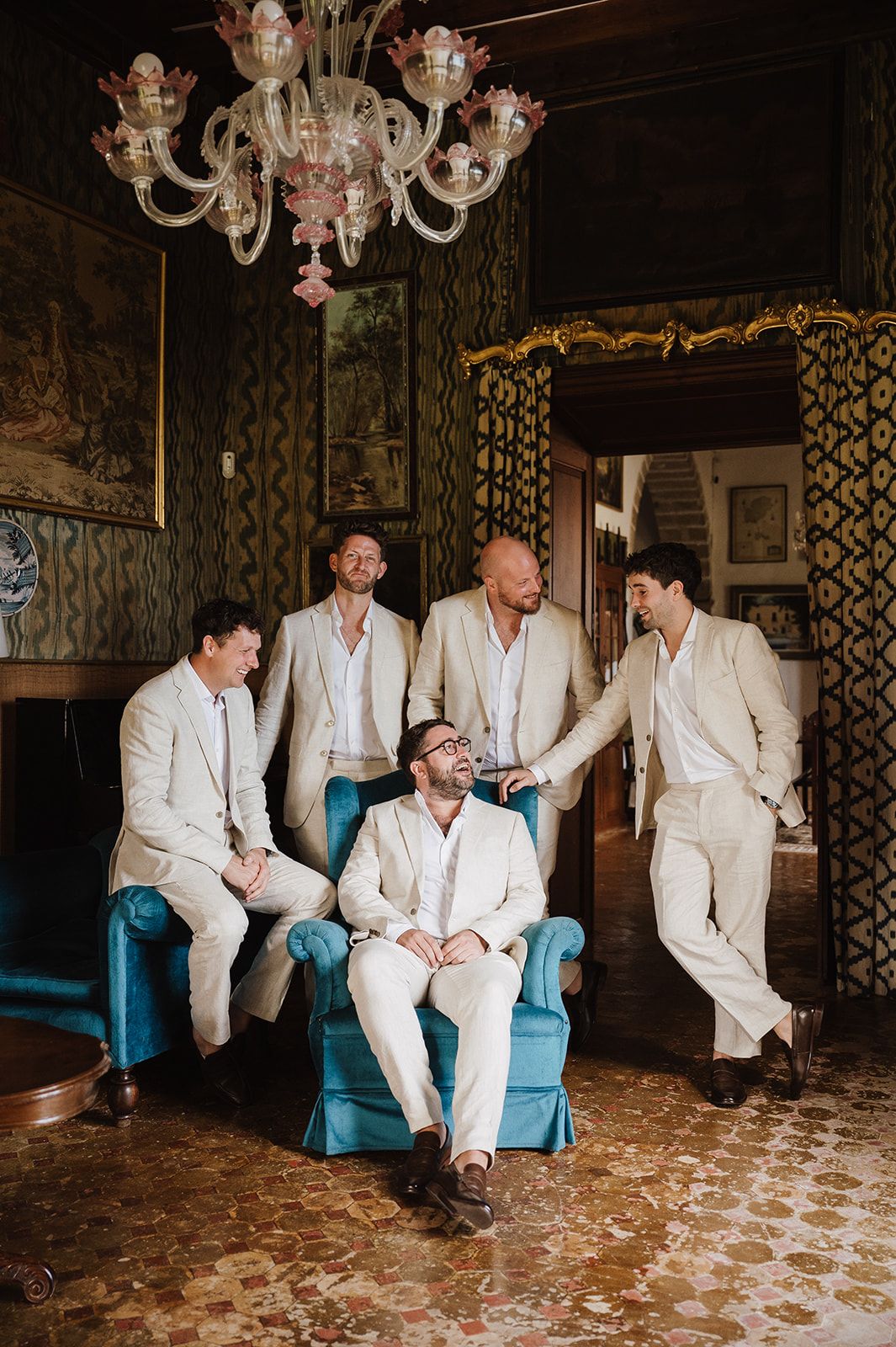 Groom sits in blue velvet chair laughing with groomsmen in cream suits inside an elegant vintage room with chandelier.