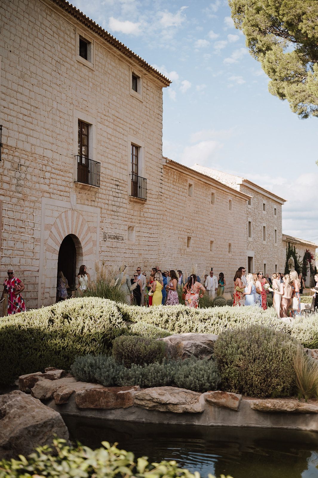 Wedding guests mingle outside a historic stone building with lush gardens and a tranquil pond in the foreground.