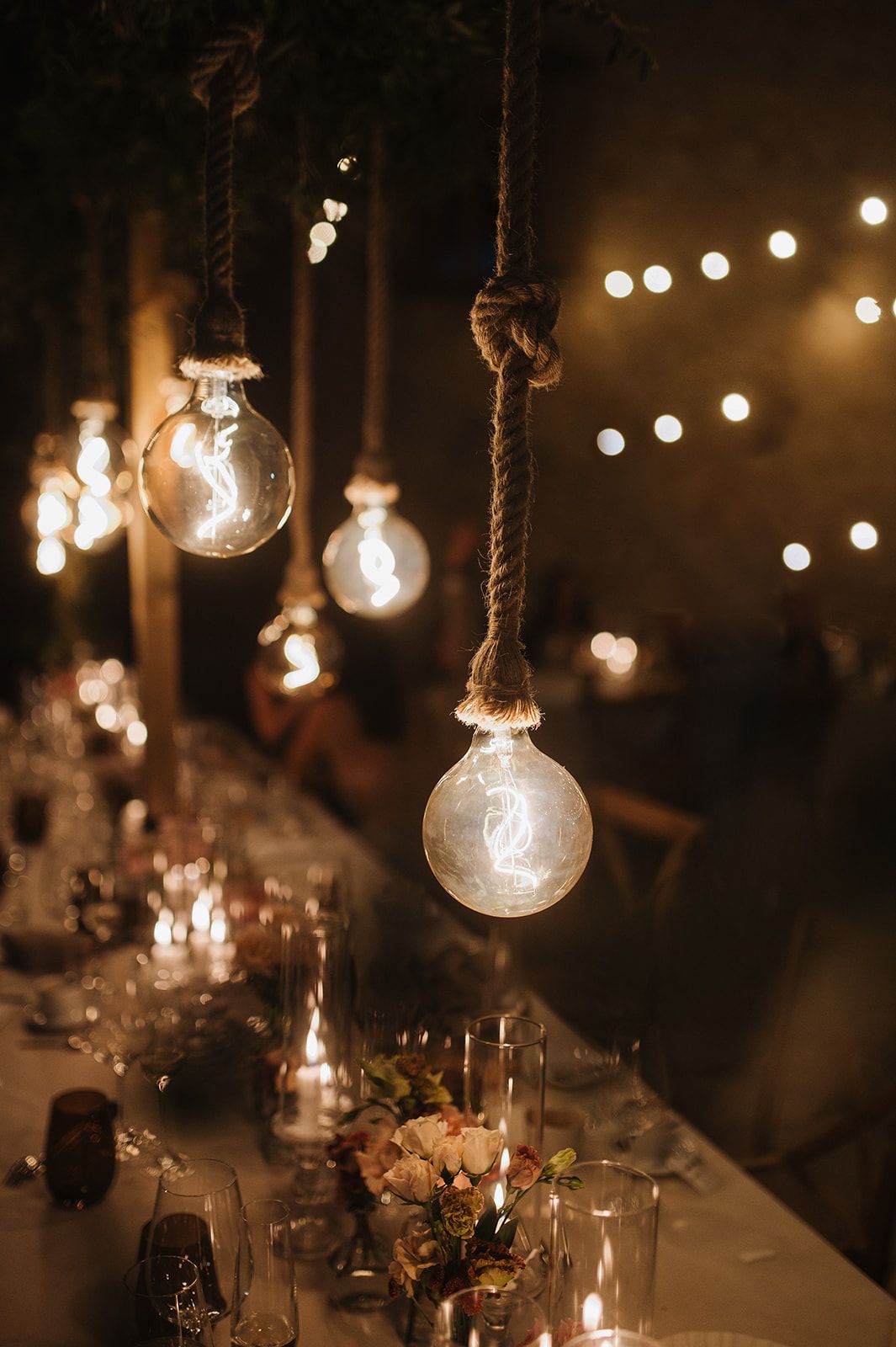 Rustic wedding reception decor featuring large Edison bulbs hanging from rope over candlelit tables.