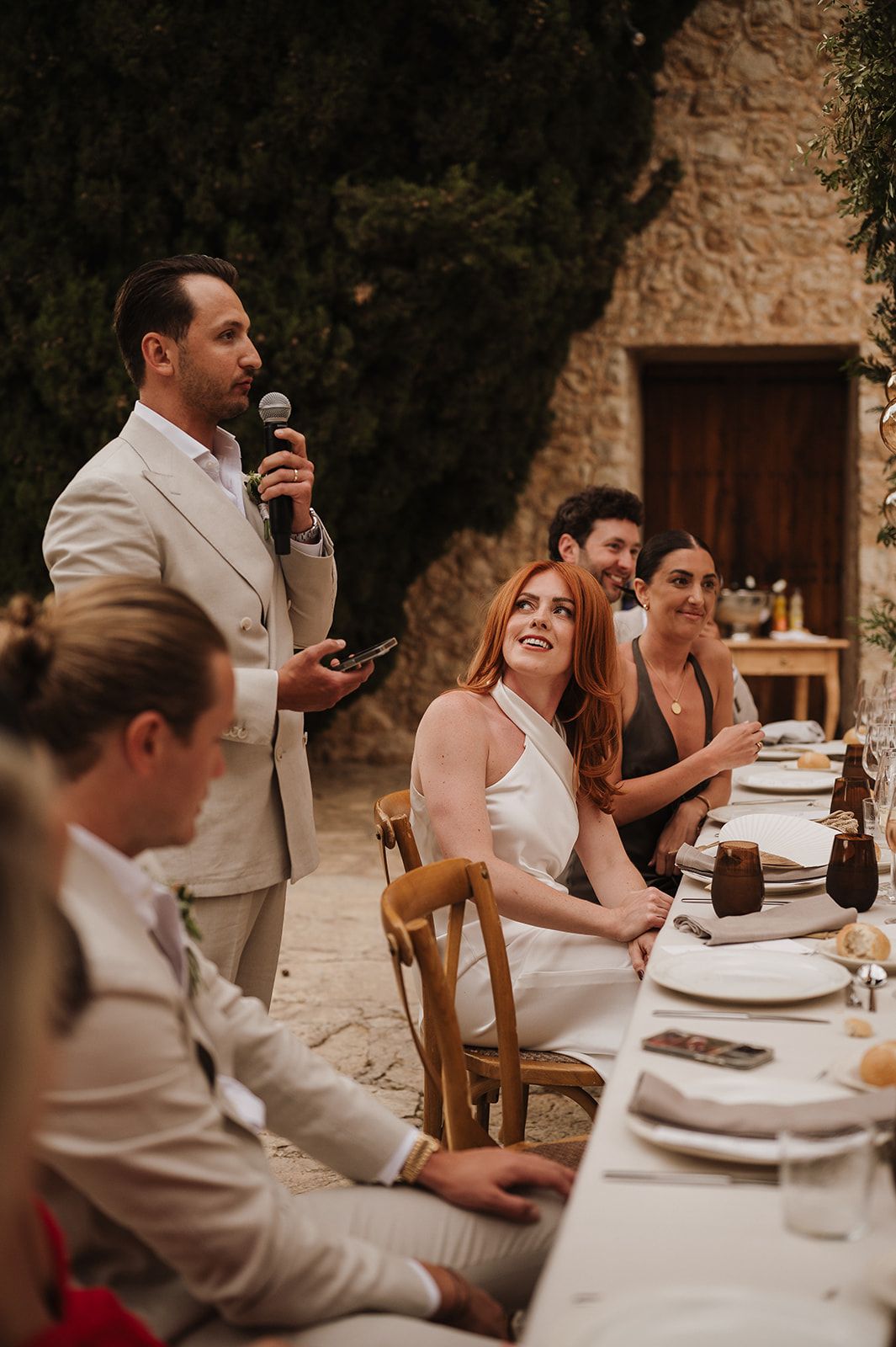 Groom speaks into microphone while bride and guests smile and listen at an elegant outdoor wedding reception.