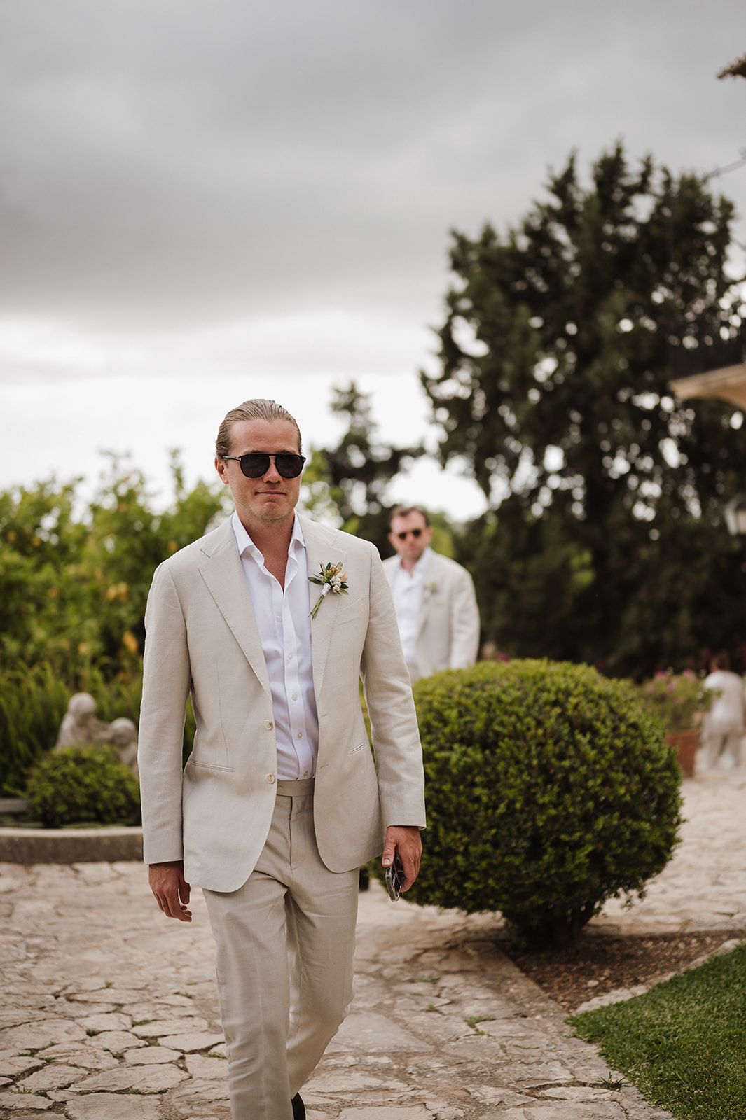 Groom in beige linen suit with boutonniere and sunglasses walks garden path at outdoor wedding.
