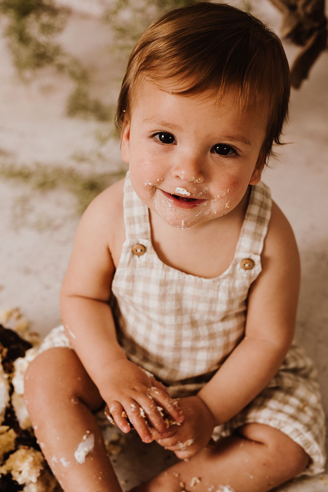 A happy baby with cake-smeared face sits in a gingham romper during a first birthday cake smash photo session.