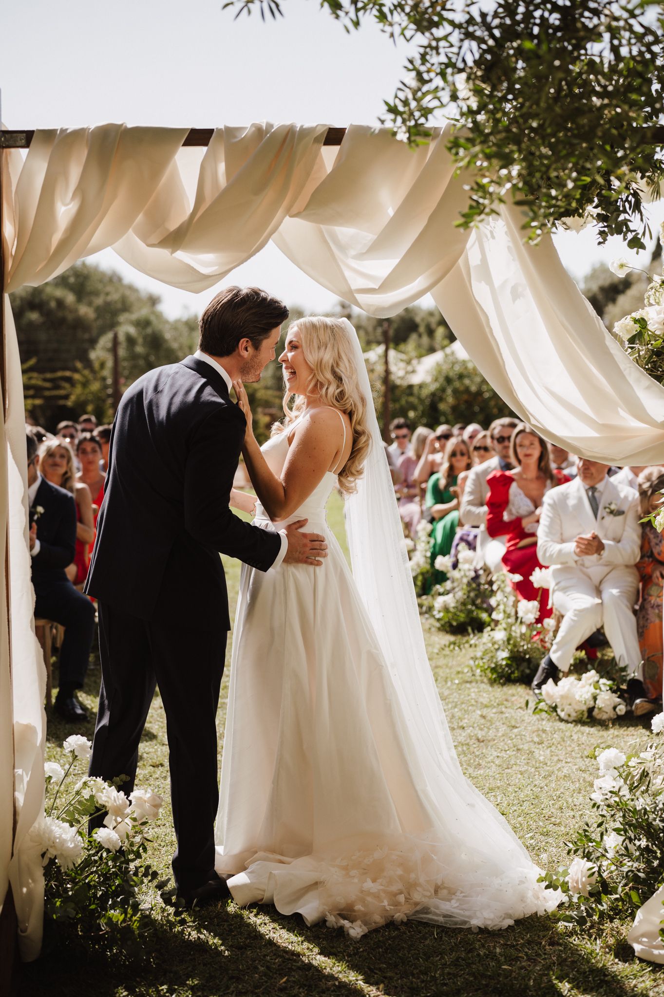 Newlyweds share first kiss under elegant draped canopy altar with white floral arrangements and seated guests.