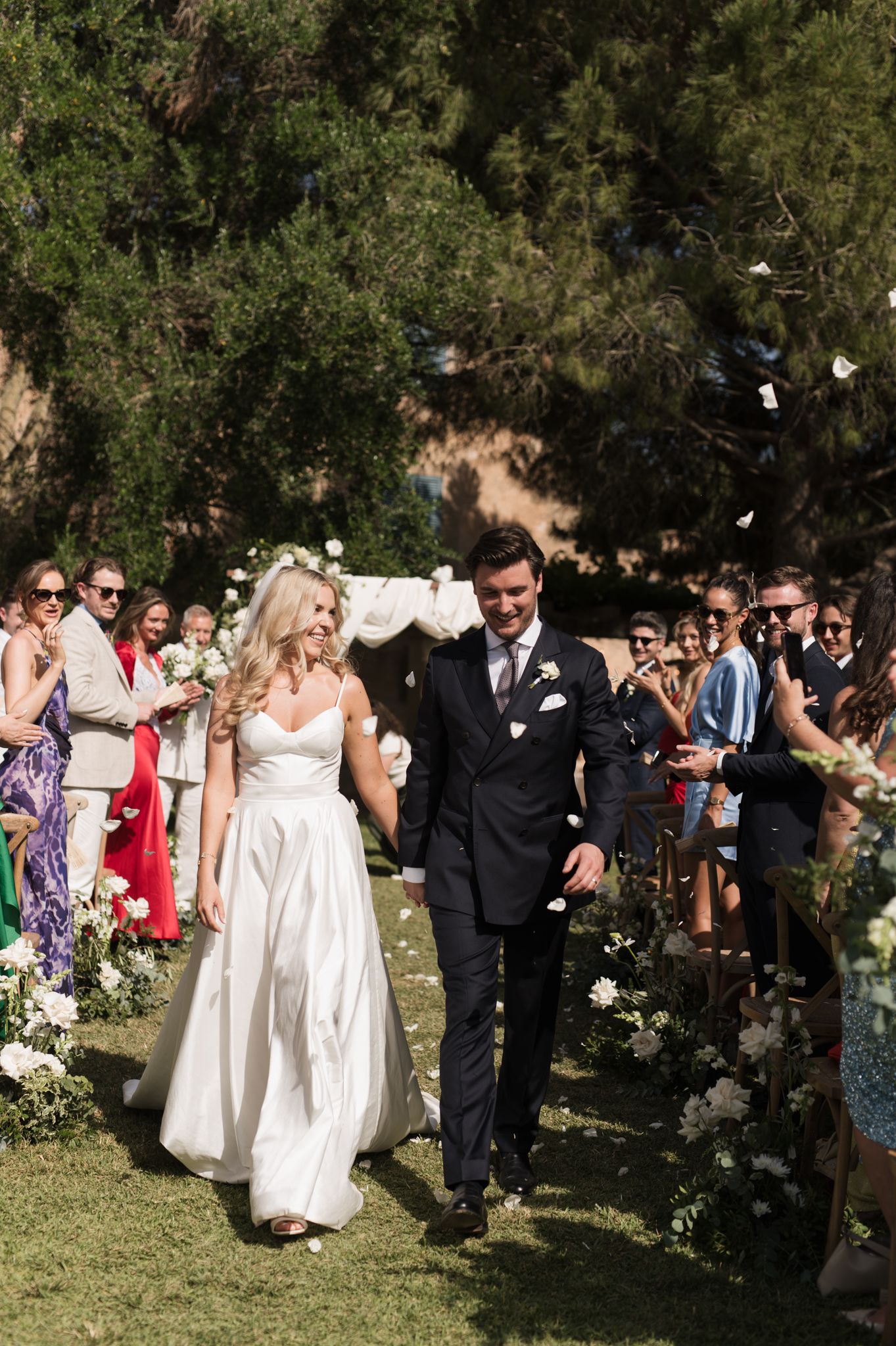 Smiling newlyweds walk down the aisle as guests toss white flower petals during outdoor wedding ceremony.