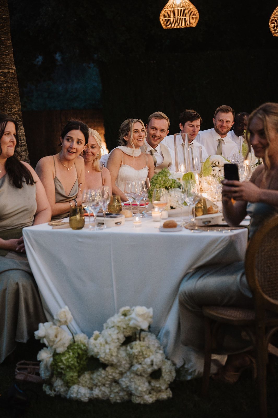 Bride and groom smile with wedding party at candlelit reception table surrounded by white floral arrangements.