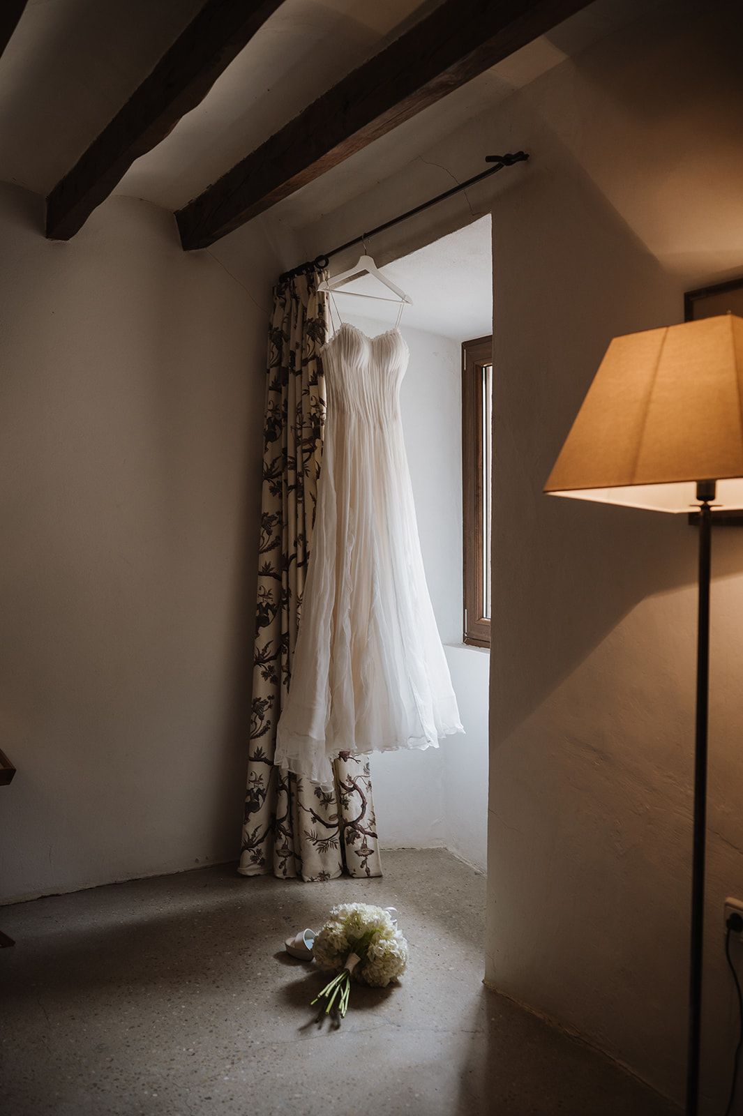 A white wedding dress hanging near a window with floral curtains, beside a floor lamp and bridal bouquet on the floor.
