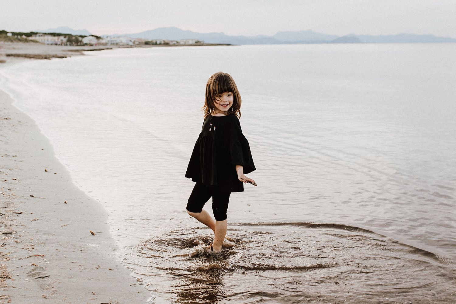 A young girl in a black dress wades barefoot in shallow ocean waves on a sandy beach.