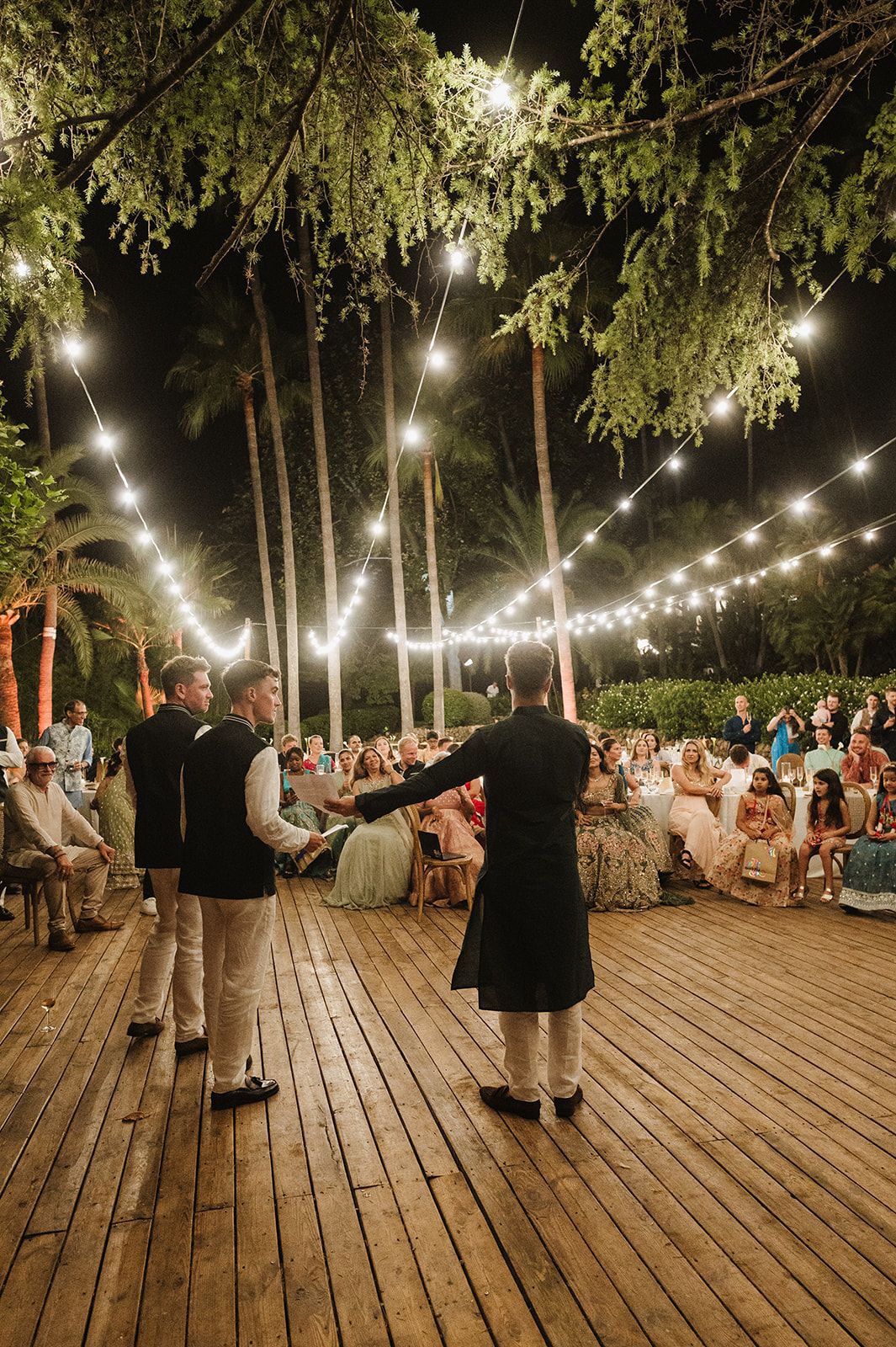 Two men perform on a wooden stage under string lights before seated wedding reception guests at night.