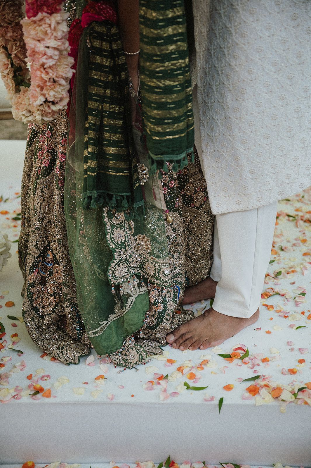 Close-up of bride in embroidered green lehenga and groom in white sherwani standing barefoot at altar.