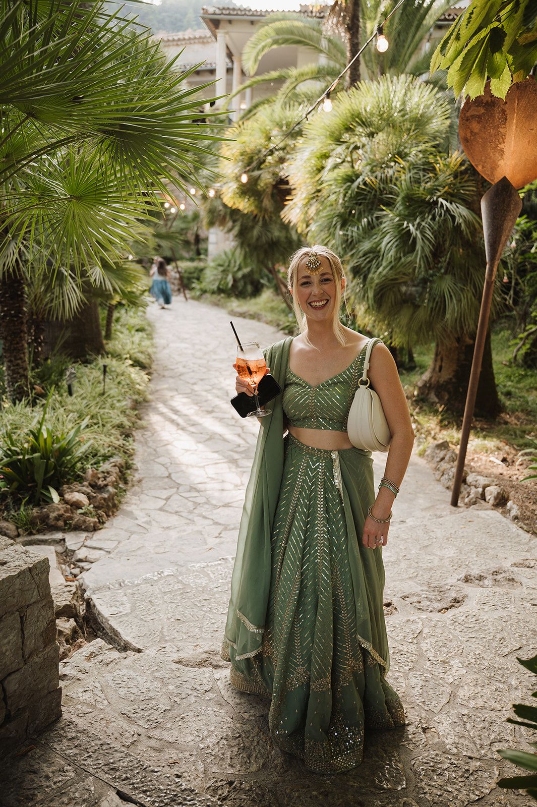 Wedding guest in green lehenga holding cocktail at outdoor Indian wedding reception.