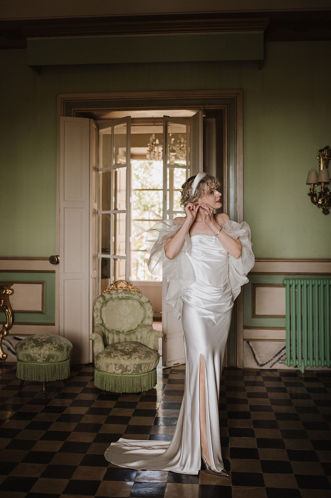 Bride in satin wedding gown with feather cape adjusts earring in elegant green vintage room with checkered floor.