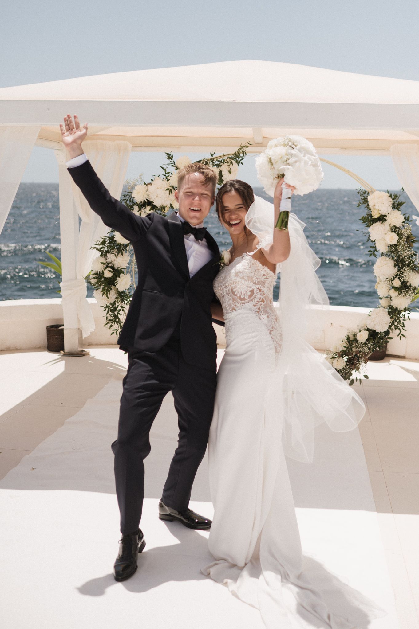 Joyful newlyweds celebrate at seaside wedding altar, groom in tuxedo raises arm as bride holds bouquet.