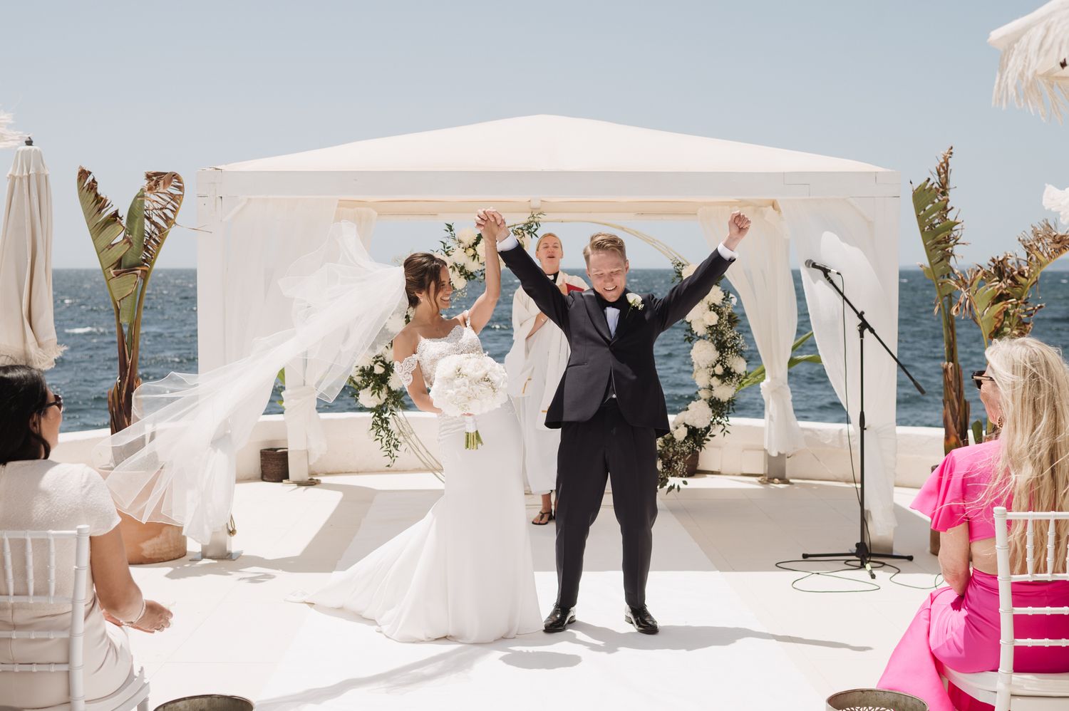 Joyful bride and groom celebrate at outdoor oceanfront wedding ceremony raising arms in triumph.