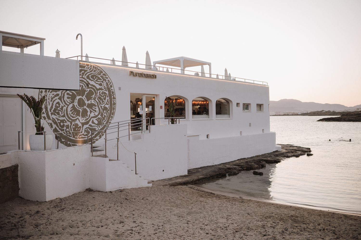 White beachside restaurant building with ornate circular decor at sunset, situated on rocky Mediterranean shore.