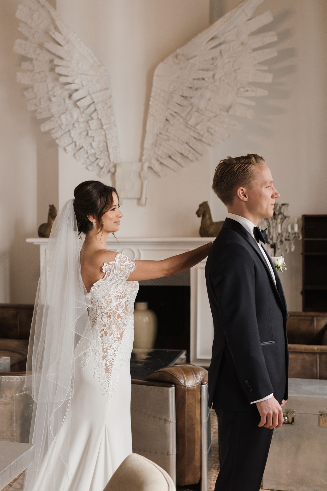 Bride in lace wedding gown touches groom's shoulder during first look with angel wings decor.