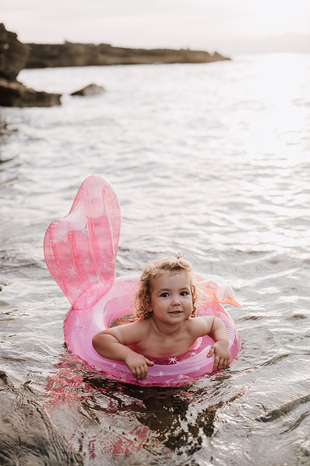 Cute toddler girl floating in a pink mermaid inflatable ring in calm ocean water.