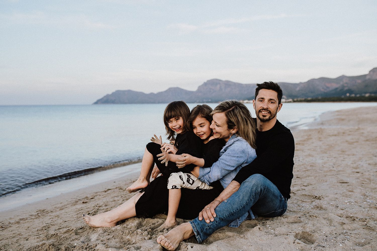 Happy family of four sitting together on a sandy beach at dusk with mountains behind.