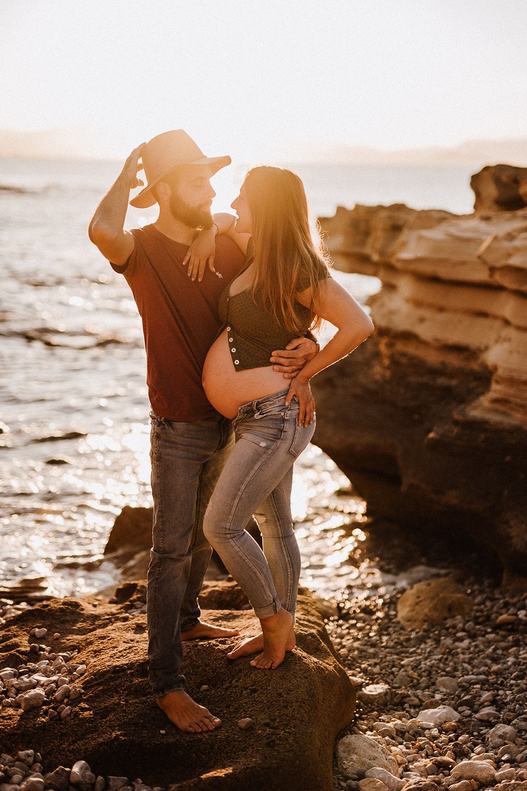 Couple embracing on rocky beach at golden sunset, pregnant woman in jeans with partner in hat by ocean.