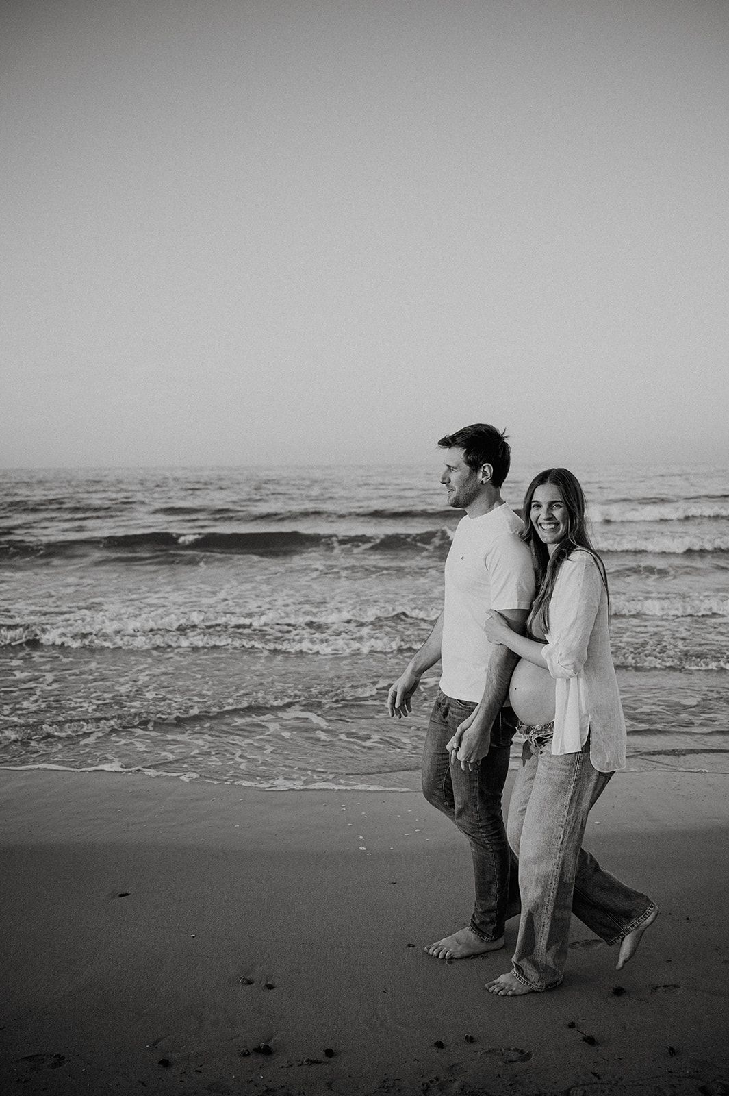 Black and white photo of a smiling pregnant couple walking barefoot on a beach by the ocean.