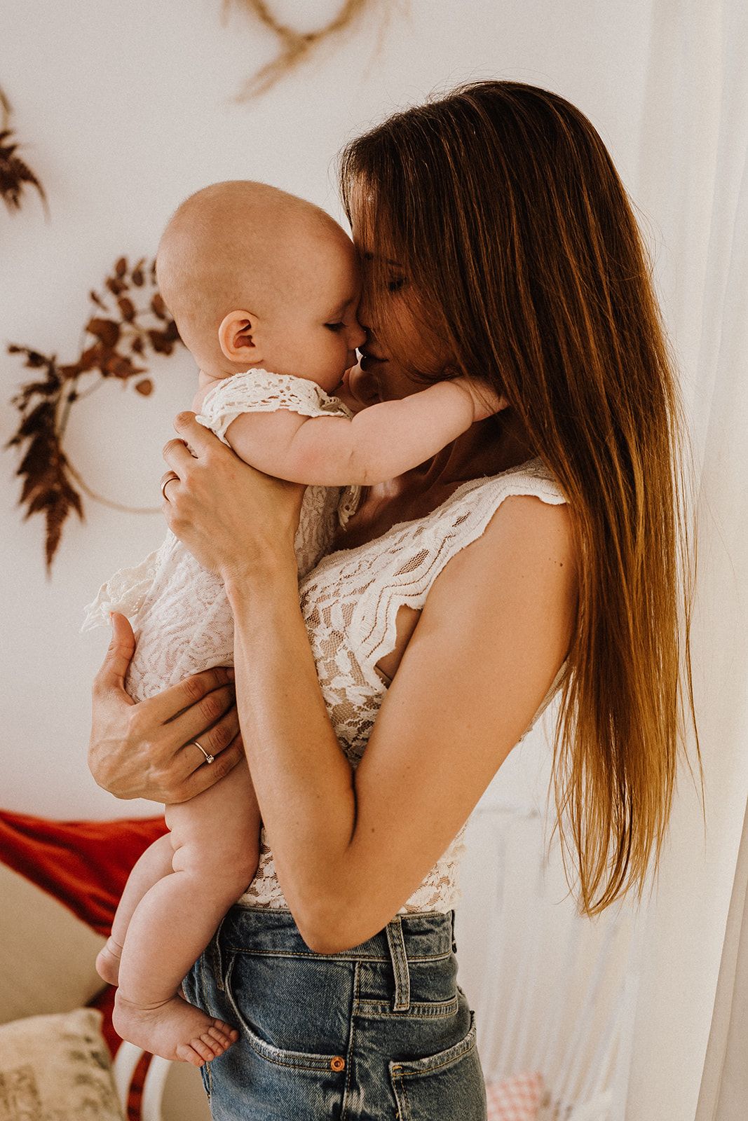A mother tenderly holds her baby close, touching foreheads in an intimate moment wearing a white lace top and jeans.