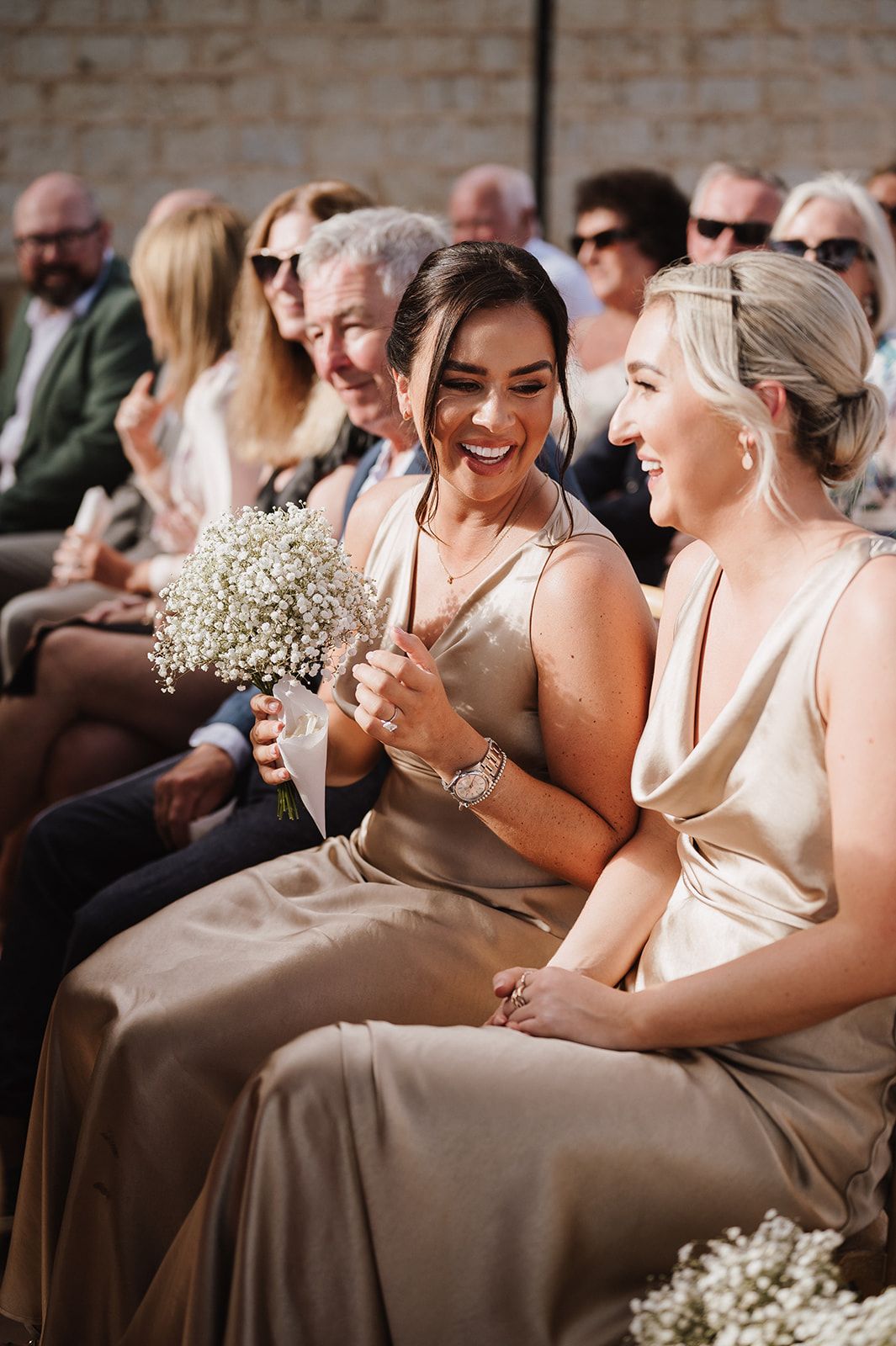 Two bridesmaids in gold satin dresses laughing joyfully while seated at an outdoor wedding ceremony.