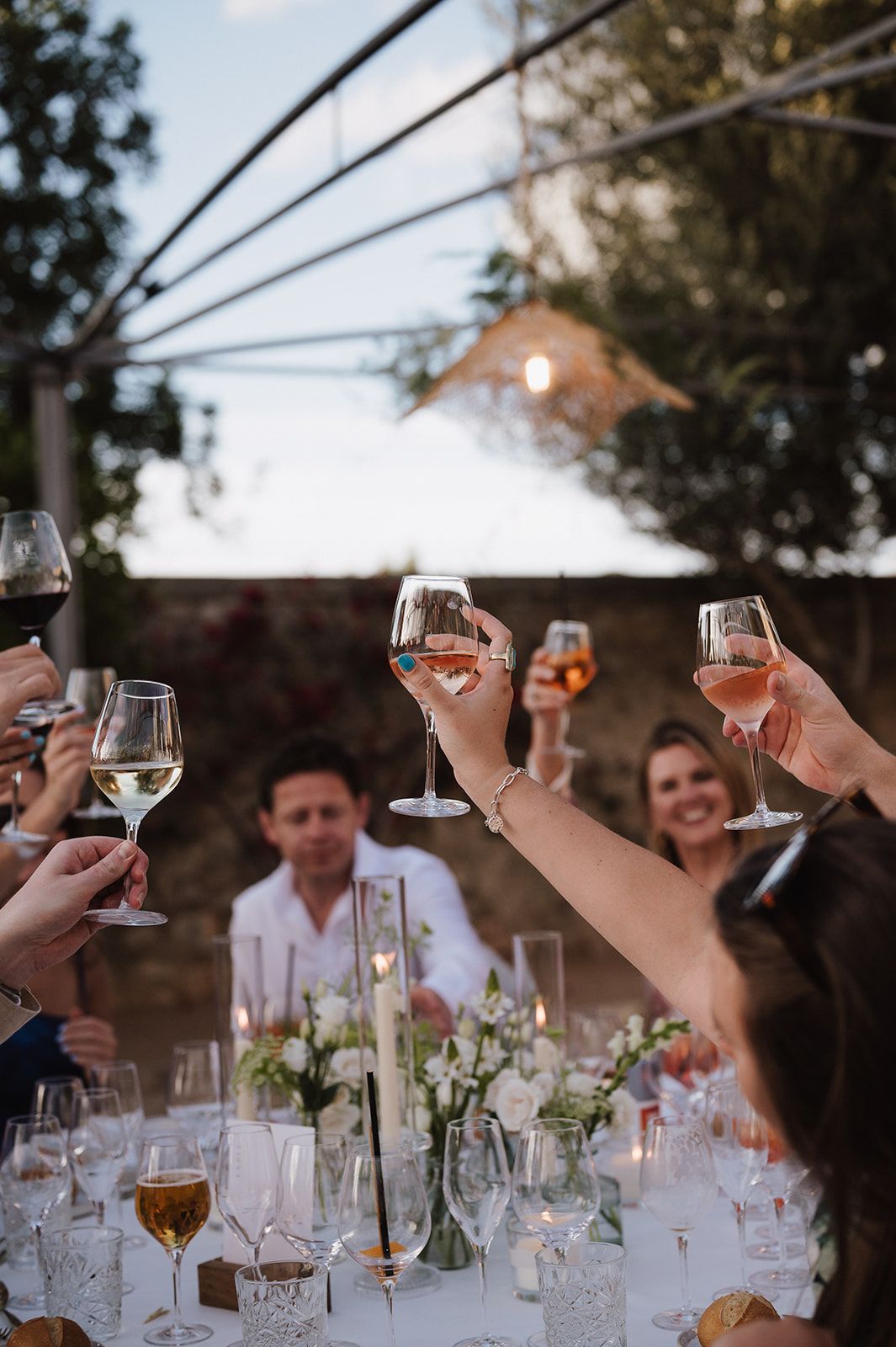 Guests raising wine glasses in a toast at an elegant outdoor wedding reception dinner with candlelit floral centerpieces.