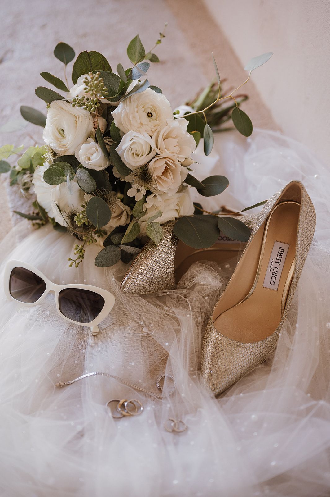 Wedding flat lay with Jimmy Choo heels, white rose bouquet, sunglasses, and rings on tulle.