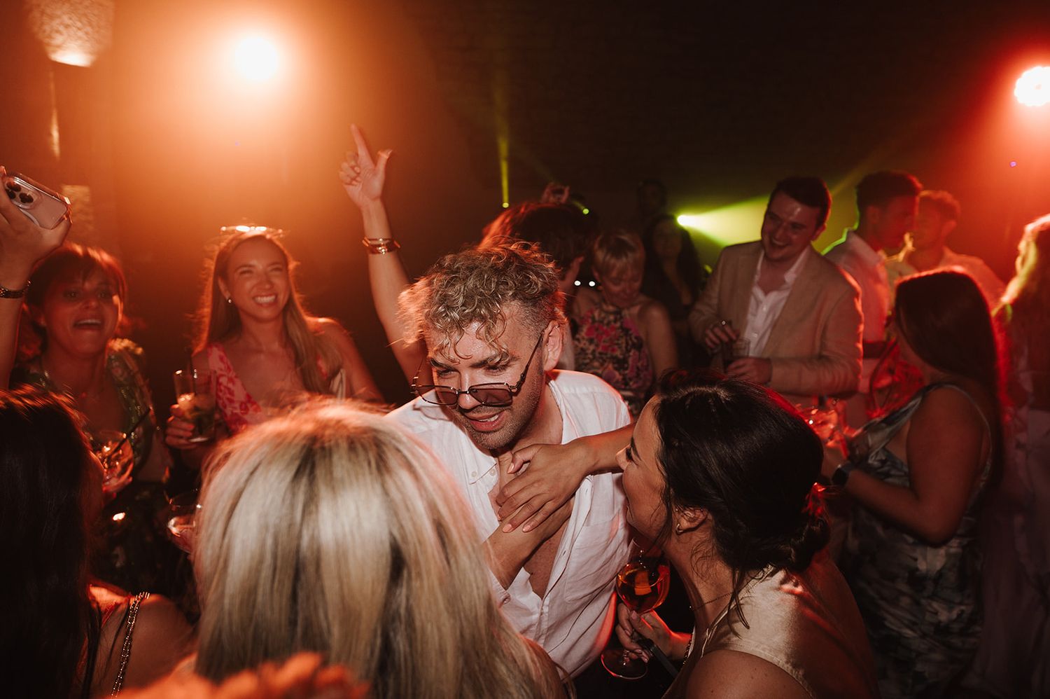 Groom dances energetically with guests under warm lights at a lively wedding party.